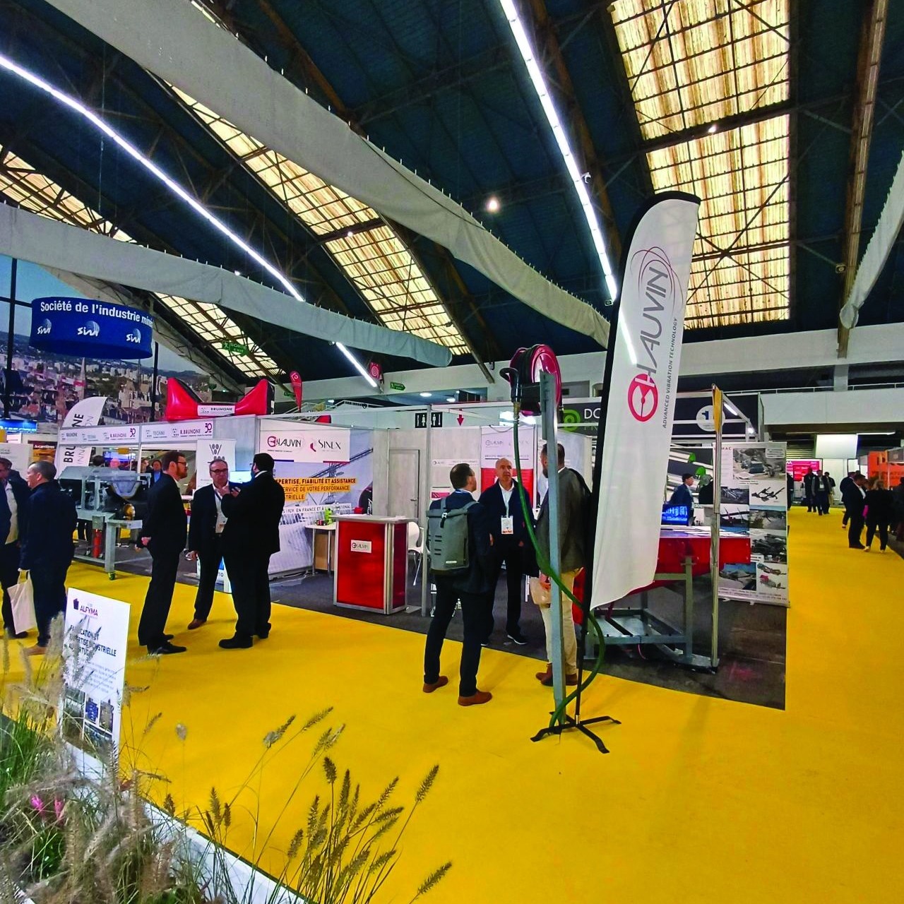 Vue d’un salon industriel avec des stands d’exposition, des représentants d’entreprises en discussion et un tapis jaune caractéristique. L’événement se déroule sous une grande structure avec un éclairage moderne.