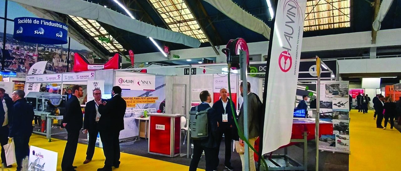 Vue d’un salon industriel avec des stands d’exposition, des représentants d’entreprises en discussion et un tapis jaune caractéristique. L’événement se déroule sous une grande structure avec un éclairage moderne.
